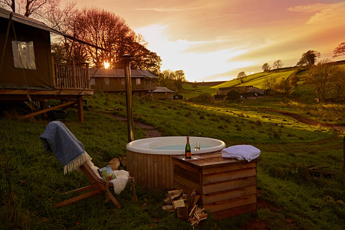 Sun setting in the sky with champagne bottle and glasses sitting next to Bovey safari tent wood-fired hot tub