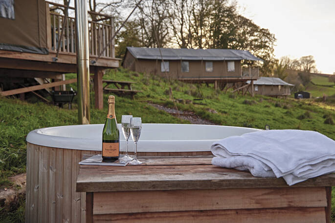 Wood-fired hot tub outside Bovey safari tent in Devon as the suns sets in the evening sky. A champagne bottle and two glasses sit on the edge 