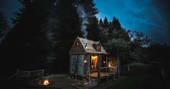 Rowan cosy cabin exterior view under a starry night sky with a roaring campfire and smoking chimney from the woodburner