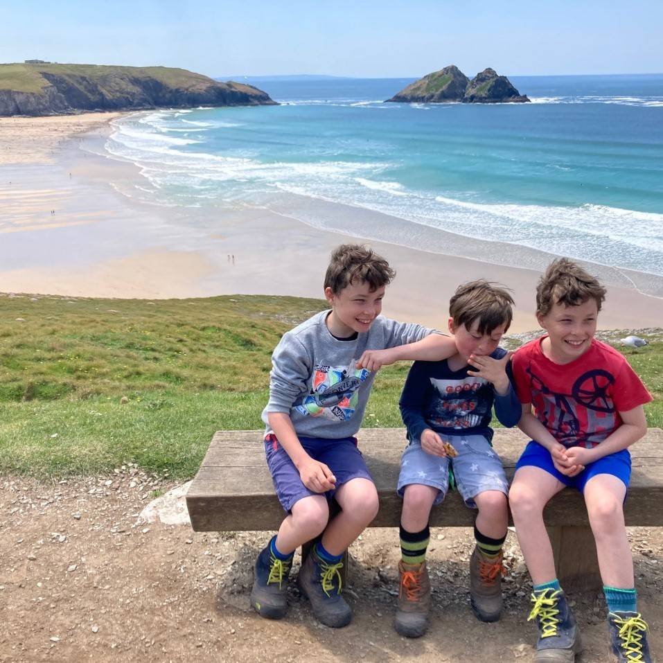 Three boys sitting on a clifftop bench above a curved bay and seascape