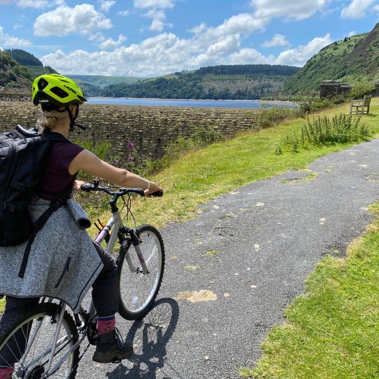 A woman on a bike with a reservoir visible in the middle distance behind a tall retaining wall