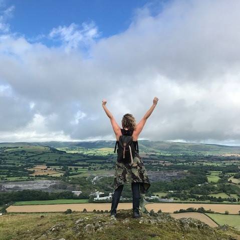 A woman standing in a triumphant pose on Stanner Rock, Powys countryside below