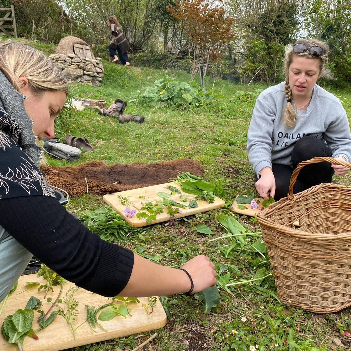 Two women kneeling in a garden with chopping boards and foraged plants